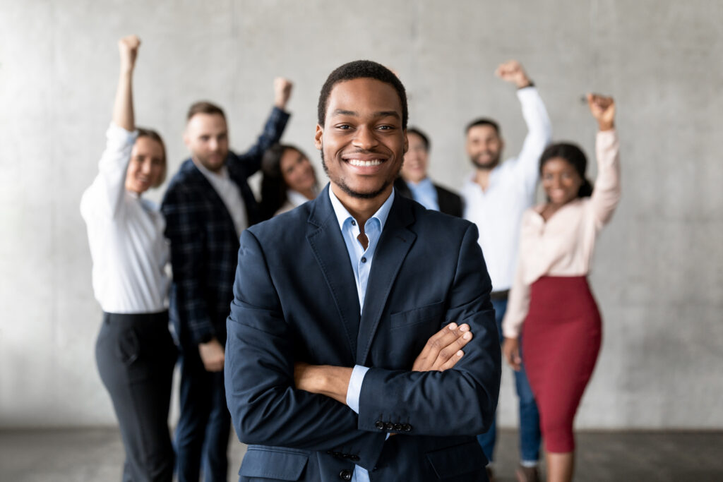 A young business professional standing confidently with his colleagues standing in the background, raising hands in celebration.